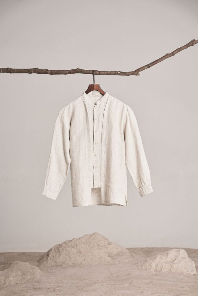 A beige shirt hangs from a branch in a minimalist studio setting with a white background.